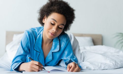 happy african american woman in pajamas lying on bed and writing on notebook